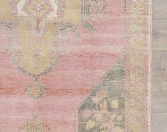 May include: A vintage rug with a pink background and a faded floral pattern in shades of beige and gray. The rug has a geometric border in shades of beige, pink, and gray.