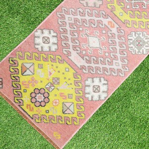 May include: A pink and yellow patterned rug with geometric designs. The rug has a fringe border and is laid out on a green grassy surface.