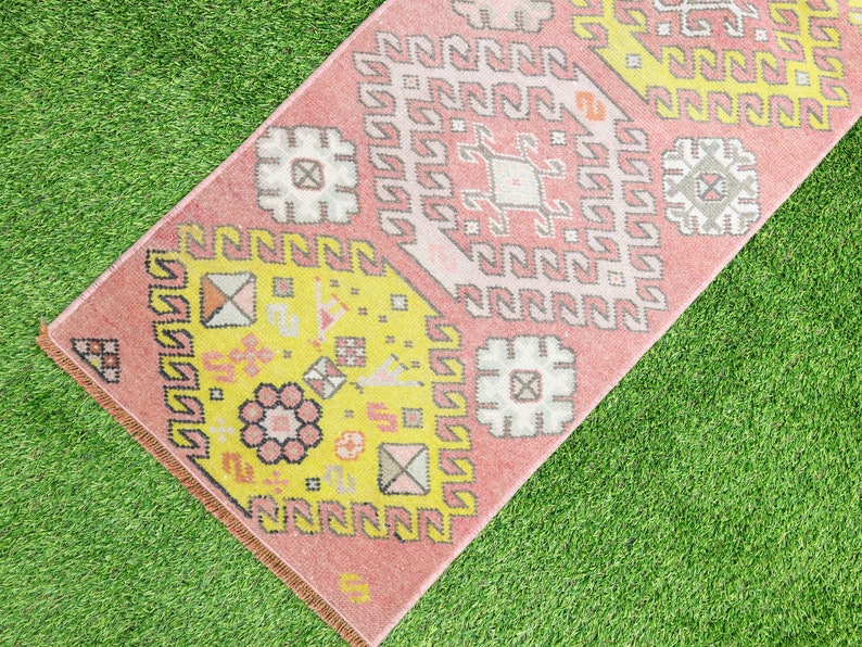 May include: A pink and yellow patterned rug with geometric designs. The rug has a fringe border and is laid out on a green grassy surface.