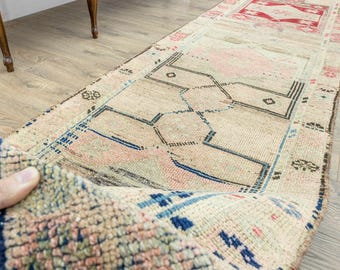 May include: A long, rectangular runner rug featuring a beige base with geometric patterns in faded red, pink, and blue. The rug displays a series of interconnected diamond and linear motifs, with a visible pile texture and a folded corner.