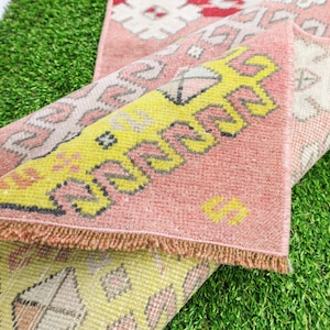 May include: Two pink and yellow patterned rugs with fringed edges are folded on top of each other. The rugs are lying on a green artificial grass surface.