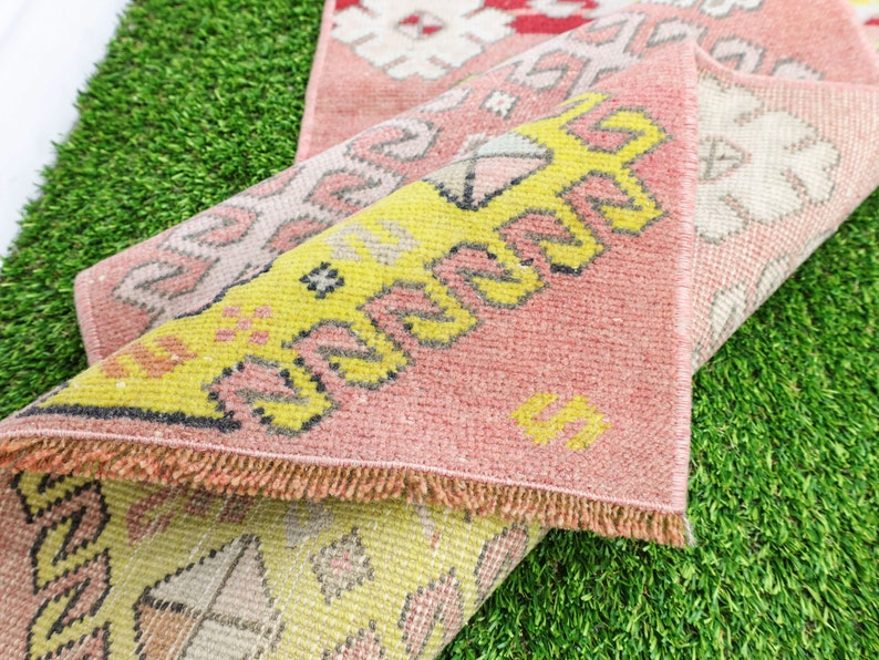 May include: Two pink and yellow patterned rugs with fringed edges are folded on top of each other. The rugs are lying on a green artificial grass surface.