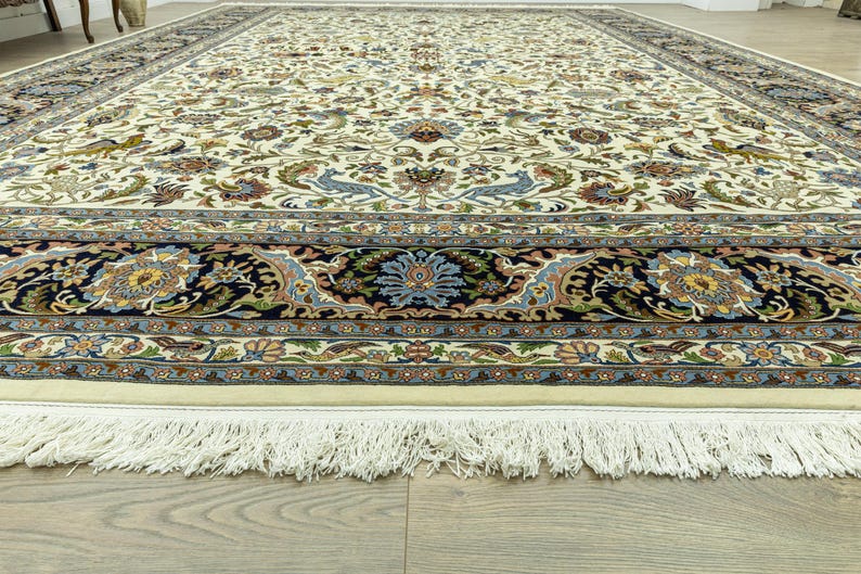 May include: An ivory-colored, rectangular area rug with an intricate floral and bird pattern. The rug features a dark blue border with floral medallions and a cream-colored fringe along the edge. The design includes shades of blue, green, and gold.