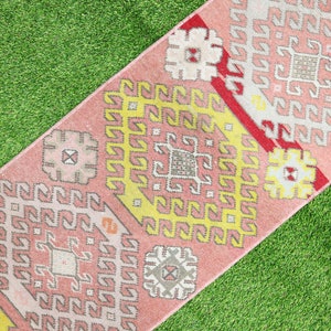 May include: A pink and yellow patterned rug with a geometric design. The rug is made of woven fabric and has a fringed edge.