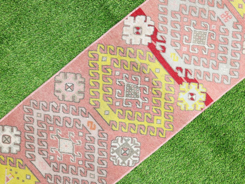 May include: A pink and yellow patterned rug with a geometric design. The rug is made of woven fabric and has a fringed edge.