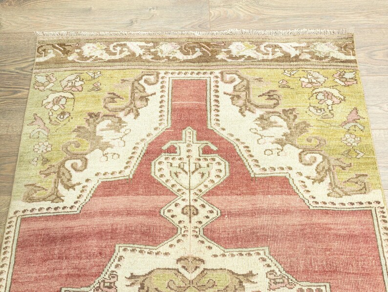 以下が含まれることがあります： ヴィンテージラグのクローズアップ。赤とクリーム色の中心的なデザインで、緑と茶色の花の模様のボーダーで囲まれています。ラグの端にはフリンジが付いています。