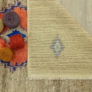 Peut inclure: Un tapis beige et orange &agrave; motifs avec une bordure bleue est partiellement visible, ainsi que plusieurs pelotes de laine jaunes, violettes, orange et rouges. Le tapis est pli&eacute;, r&eacute;v&eacute;lant un motif ray&eacute;.