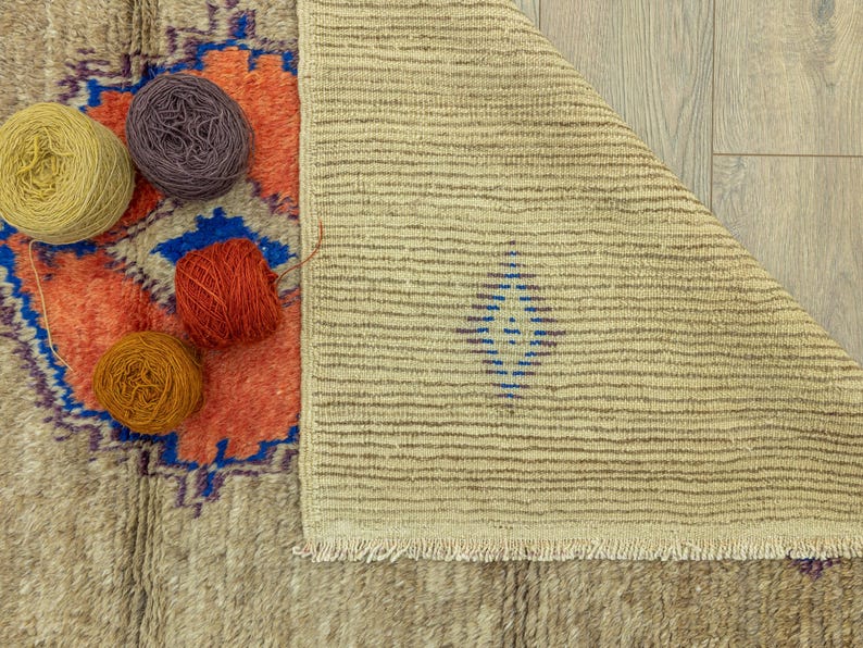 Peut inclure: Un tapis beige et orange &agrave; motifs avec une bordure bleue est partiellement visible, ainsi que plusieurs pelotes de laine jaunes, violettes, orange et rouges. Le tapis est pli&eacute;, r&eacute;v&eacute;lant un motif ray&eacute;.