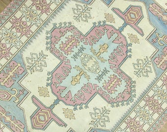 May include: A vintage rug with a central medallion design in pastel colors. The rug features a cream-colored background with a central pattern in light blue, pink, and yellow. The border has geometric patterns in pink and cream.