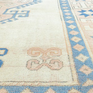 May include: A vintage rug with a cream background and a blue and pink geometric pattern. The rug has a faded look and is made of a soft, woven material.