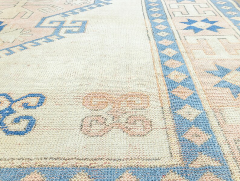 May include: A vintage rug with a cream background and a blue and pink geometric pattern. The rug has a faded look and is made of a soft, woven material.