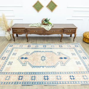 May include: A cream-coloured rug with a blue and pink geometric pattern. The rug has a central medallion with a repeating design of stylised flowers and leaves. The rug is laid out on a wooden floor in a room with white walls and a wooden console table.