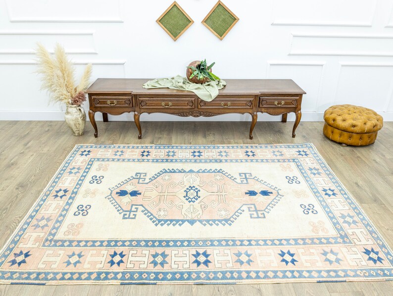 May include: A cream-coloured rug with a blue and pink geometric pattern. The rug has a central medallion with a repeating design of stylised flowers and leaves. The rug is laid out on a wooden floor in a room with white walls and a wooden console table.