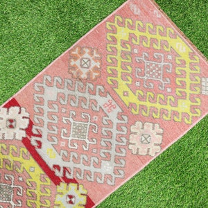 May include: A pink and yellow patterned rug with a geometric design. The rug has a fringe border and is laid out on a green grassy surface.