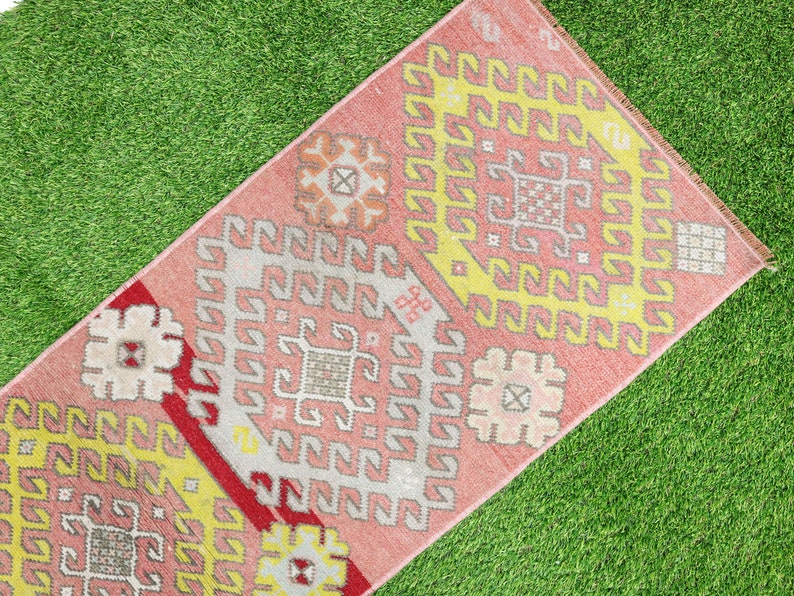 May include: A pink and yellow patterned rug with a geometric design. The rug has a fringe border and is laid out on a green grassy surface.