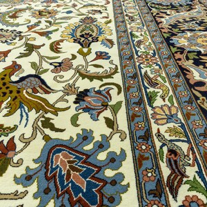 May include: Close-up of a detailed Persian rug with a cream-colored background. The rug features intricate floral and bird patterns in shades of blue, green, brown, and red. The border has a blue and cream floral design.