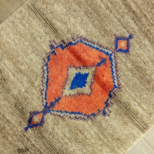 Peut inclure: Tapis beige avec un motif g&eacute;om&eacute;trique central orange, bleu et beige. Le tapis a une surface textur&eacute;e et un motif en forme de losange. Le motif est soulign&eacute; en bleu et comporte un losange au centre.