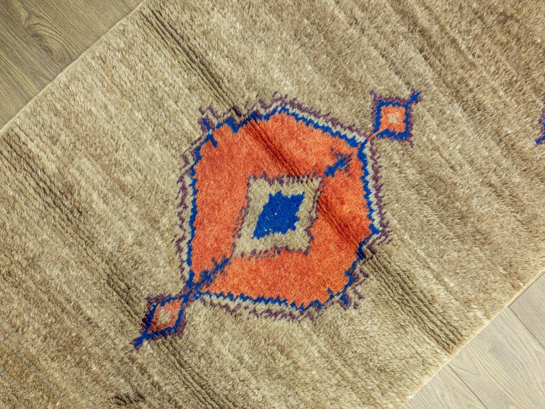 Peut inclure: Tapis beige avec un motif g&eacute;om&eacute;trique central orange, bleu et beige. Le tapis a une surface textur&eacute;e et un motif en forme de losange. Le motif est soulign&eacute; en bleu et comporte un losange au centre.