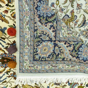 May include: An ornate, cream-colored rug with a detailed floral and animal pattern. The rug features a navy blue border with intricate designs and a fringed edge. Several balls of yarn in yellow, red, and purple are placed on the rug.