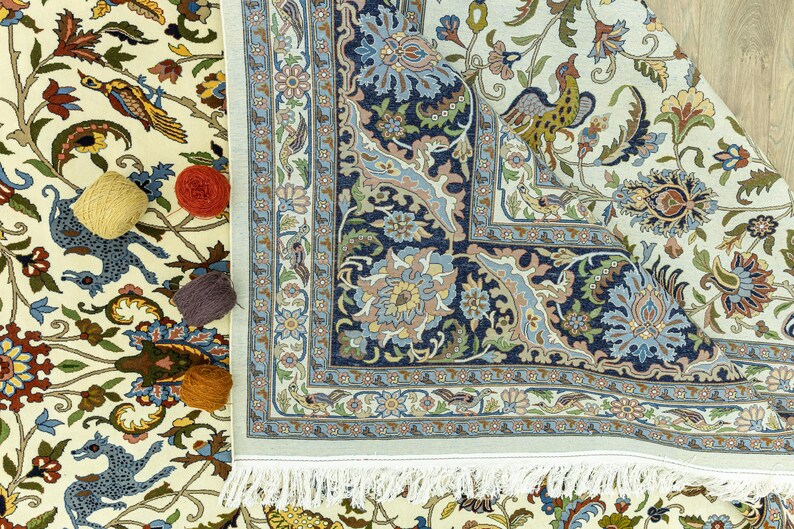May include: An ornate, cream-colored rug with a detailed floral and animal pattern. The rug features a navy blue border with intricate designs and a fringed edge. Several balls of yarn in yellow, red, and purple are placed on the rug.