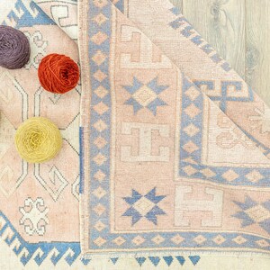 May include: A close-up of a light pink and blue patterned rug with a fringed border. Four balls of yarn in different colours are placed on the rug.
