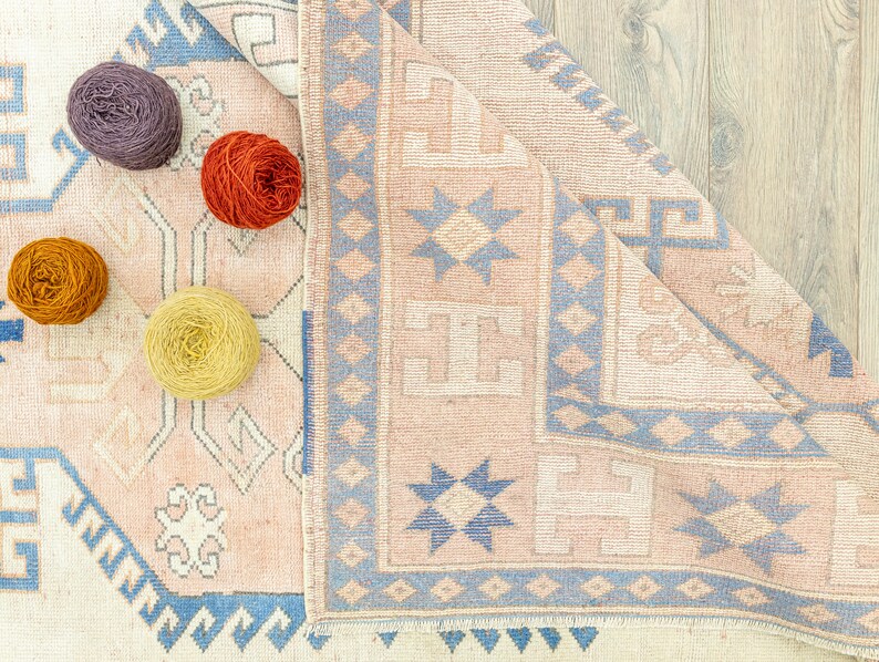 May include: A close-up of a light pink and blue patterned rug with a fringed border. Four balls of yarn in different colours are placed on the rug.