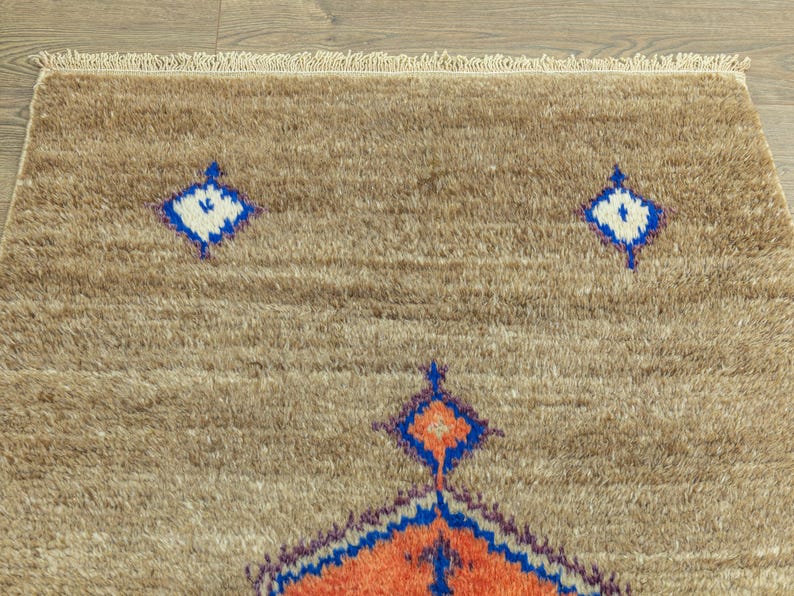 Peut inclure: Gros plan sur un tapis beige avec une surface textur&eacute;e et une bordure &agrave; franges. Le tapis pr&eacute;sente un motif r&eacute;p&eacute;titif de losanges bleus, blancs et oranges. Le tapis est fait d'une mati&egrave;re douce et moelleuse.
