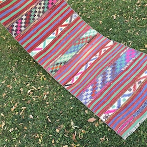 May include: A colourful striped rug with geometric patterns in shades of red, green, blue, and white. The rug is woven with a fringe on the ends and is laid out on a grassy surface.