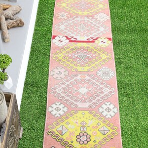 May include: A pink and yellow patterned runner rug with a geometric design. The rug has a fringed edge and is laid out on a green artificial grass surface.