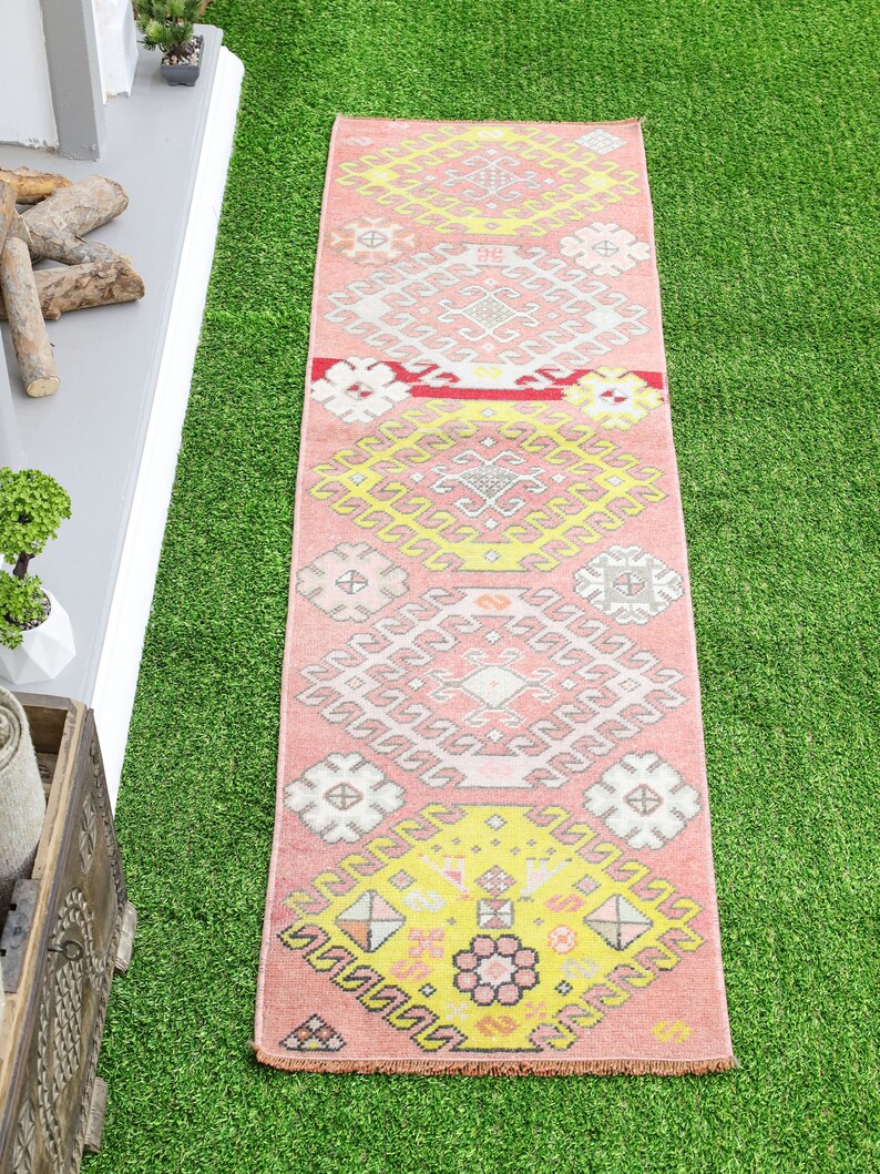 May include: A pink and yellow patterned runner rug with a geometric design. The rug has a fringed edge and is laid out on a green artificial grass surface.