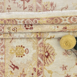 May include: A beige rug with a pink and red floral pattern. The rug has a fringe border and is folded in half. There are three balls of yarn in the bottom right corner of the image: yellow, green, and red.