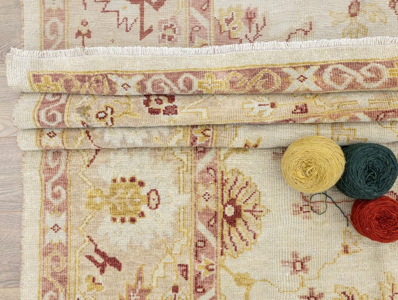 May include: A beige rug with a pink and red floral pattern. The rug has a fringe border and is folded in half. There are three balls of yarn in the bottom right corner of the image: yellow, green, and red.