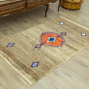 Peut inclure: Un tapis rectangulaire beige avec un motif g&eacute;om&eacute;trique central orange, bleu et blanc. Le tapis a une frange sur les petits c&ocirc;t&eacute;s et est pos&eacute; sur un parquet. Le motif du tapis comprend des losanges.