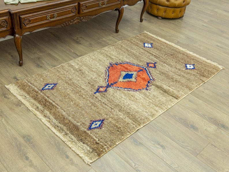 Peut inclure: Un tapis rectangulaire beige avec un motif g&eacute;om&eacute;trique central orange, bleu et blanc. Le tapis a une frange sur les petits c&ocirc;t&eacute;s et est pos&eacute; sur un parquet. Le motif du tapis comprend des losanges.