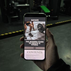 May include: A smartphone displays the cover of a fitness guide titled "ELEVATE: The Modern Guide to Health & Fitness." The image on the screen shows a person lifting a barbell in a gym setting. The phone is held in front of a blurred gym background.