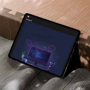 Pode incluir: Um tablet preto exibindo uma tela azul escura com o texto "Dev Brain OS". A tela mostra um monitor de computador com código, uma planta e formas geométricas. O tablet está sobre um cobertor cinza ao lado de uma superfície de madeira.