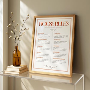 Könnte beinhalten: Ein gerahmtes Schild mit orangefarbenem Text "HOUSE RULES". Das Schild listet Regeln für ein Anwesen auf, einschließlich Geräte, Klimaanlage und Ruhezeiten. Eine braune Glasvase mit Blumen steht neben einem Stapel Bücher.