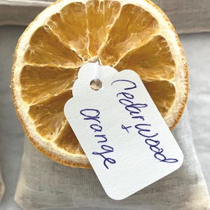 May include: A dried orange slice with a white tag that reads "Cedar Wood & Orange" in blue ink. The orange slice is on top of a small, light-colored fabric bag.