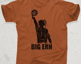 May include: An orange t-shirt featuring a black silhouette of a person holding a floral design. The text "BIG ERN" is printed in a distressed font below the silhouette. The shirt has short sleeves and a crew neck.