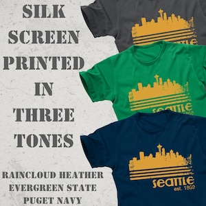 May include: Three t-shirts in raincloud heather, evergreen state, and puget navy. Each shirt features a gold-colored Seattle skyline graphic with the text "Seattle est. 1869". The image also includes the text "SILK SCREEN PRINTED IN THREE TONES".