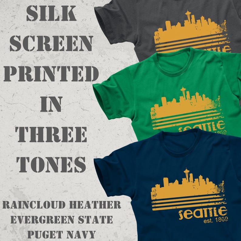 May include: Three t-shirts in raincloud heather, evergreen state, and puget navy. Each shirt features a gold-colored Seattle skyline graphic with the text "Seattle est. 1869". The image also includes the text "SILK SCREEN PRINTED IN THREE TONES".