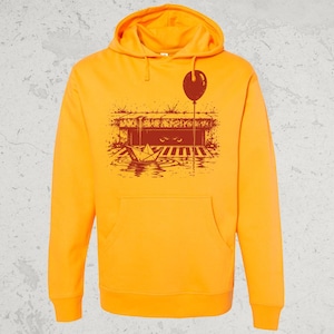 Könnte beinhalten: Ein leuchtend orangefarbener Kapuzenpullover mit einem dunkelroten Grafikdruck auf der Brust. Das Motiv zeigt ein Papierboot in einer Pfütze vor einem Gully, einen daran befestigten Luftballon sowie zwei Augen, die aus der Dunkelheit hervorschauen.