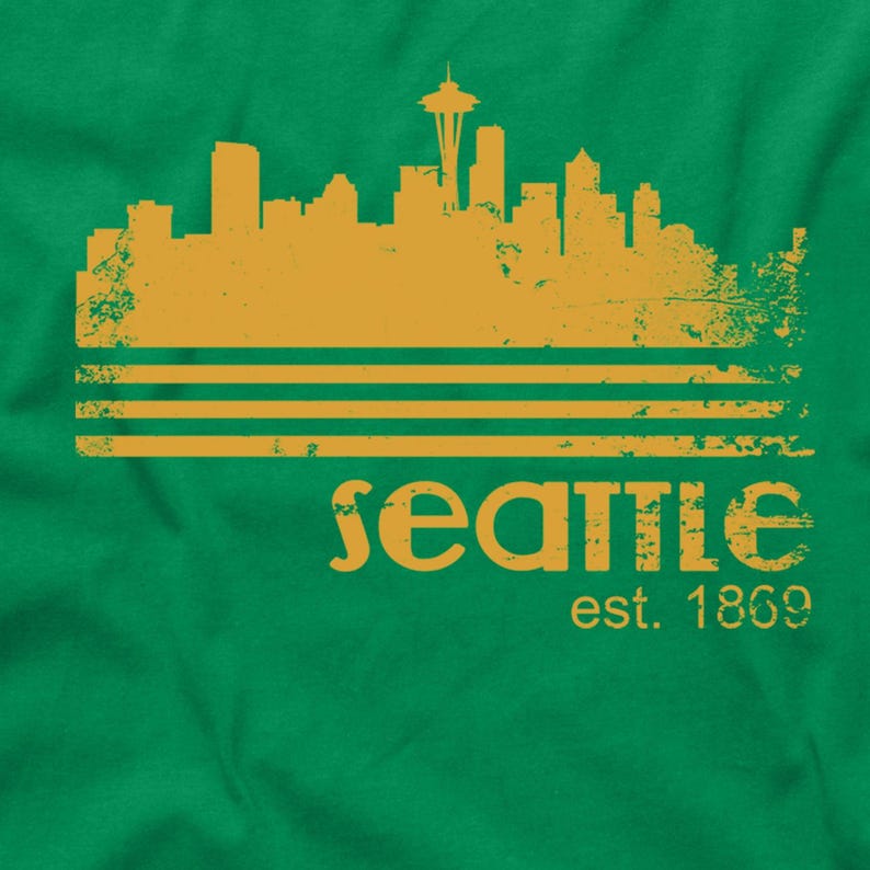 May include: A green t-shirt featuring a distressed gold design of the Seattle skyline, three horizontal lines, and the word "Seattle" with "est. 1869" below. The Space Needle is prominently displayed.