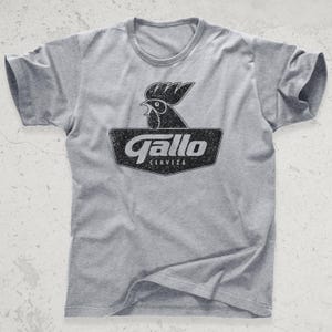 May include: A grey t-shirt with a black and white rooster graphic and the text "Gallo Cerveza". The design is distressed. The shirt has a crew neck and short sleeves.