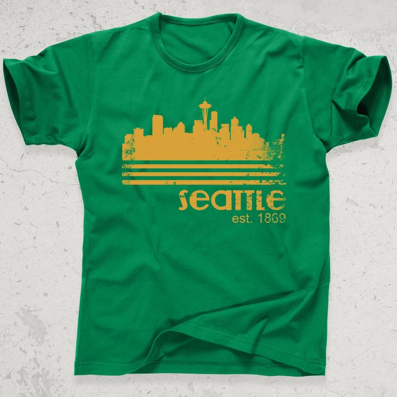 May include: Green t-shirt featuring a gold-colored Seattle skyline design with the Space Needle. The word "Seattle" is printed below the skyline, along with "est. 1869". The shirt is made of a soft material.