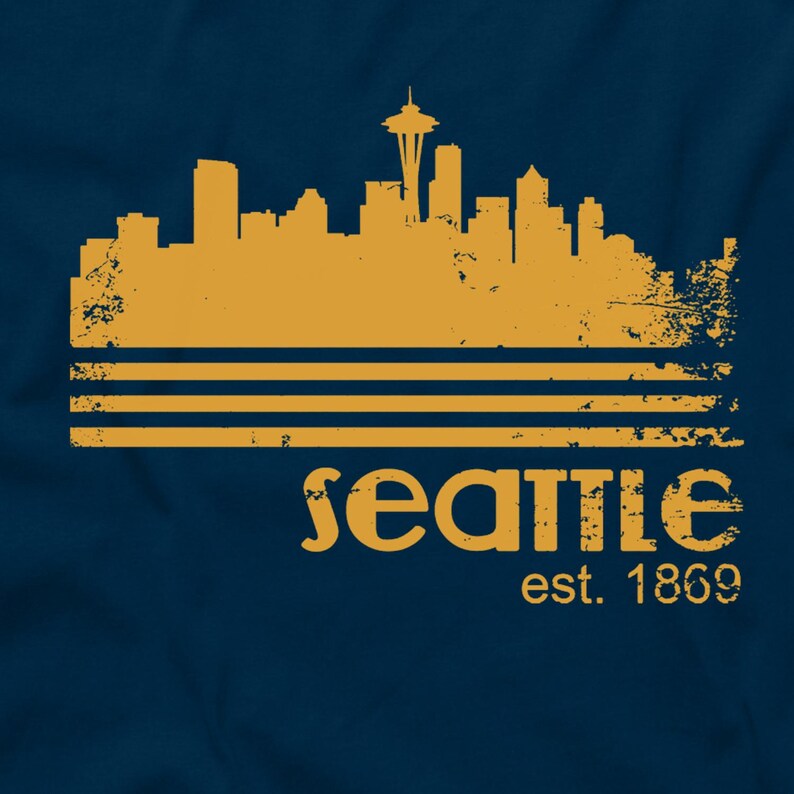 May include: Navy blue t-shirt featuring a distressed gold-colored Seattle skyline graphic. The design includes the Space Needle and the text "Seattle est. 1869". The graphic is above three horizontal lines.