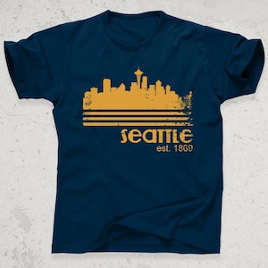 May include: Navy blue t-shirt featuring a gold-colored Seattle skyline graphic with the Space Needle. The word "Seattle" is printed below the skyline, along with "est. 1869". A casual tee for fans of the city.