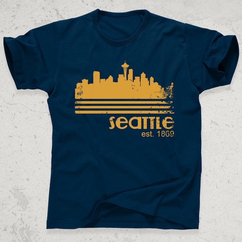 May include: Navy blue t-shirt featuring a gold-colored Seattle skyline graphic with the Space Needle. The word "Seattle" is printed below the skyline, along with "est. 1869". A casual tee for fans of the city.