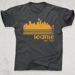 May include: Dark gray t-shirt featuring a distressed gold skyline graphic of Seattle, Washington, with the Space Needle. The word "Seattle" and the year "est. 1869" are printed below the skyline in gold.