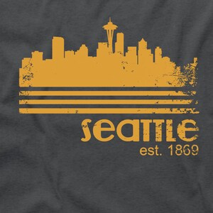 May include: A dark gray t-shirt featuring a distressed gold-colored graphic of the Seattle skyline, with the Space Needle prominent. Below the skyline are three horizontal lines and the word "Seattle" with "est. 1869" underneath.